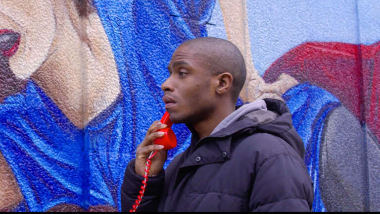 Een man neemt de rode telefoon op die geplaatst is naast de muurschildering van "An emergency call for respect". Hij luistert naar een verhaal van iemand die tijdens een interventie te maken had met agressie.