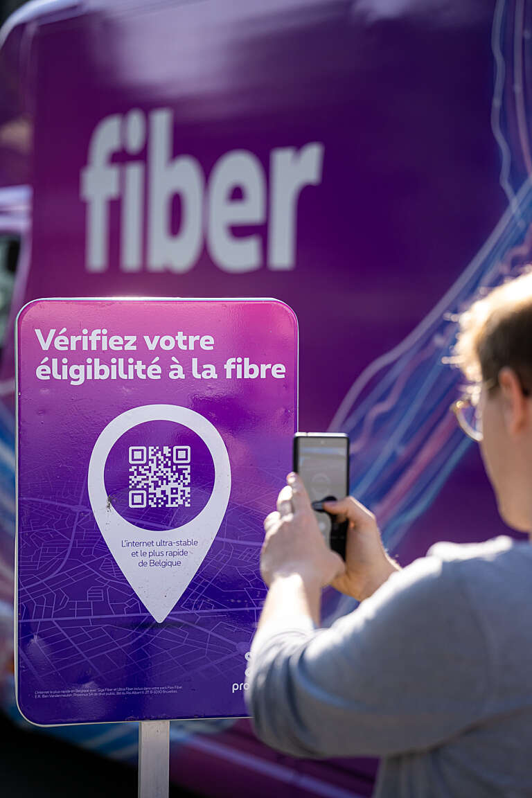 Personne scannant un QR code sur un panneau indiquant "Vérifiez votre éligibilité à la fibre", devant le van Proximus..