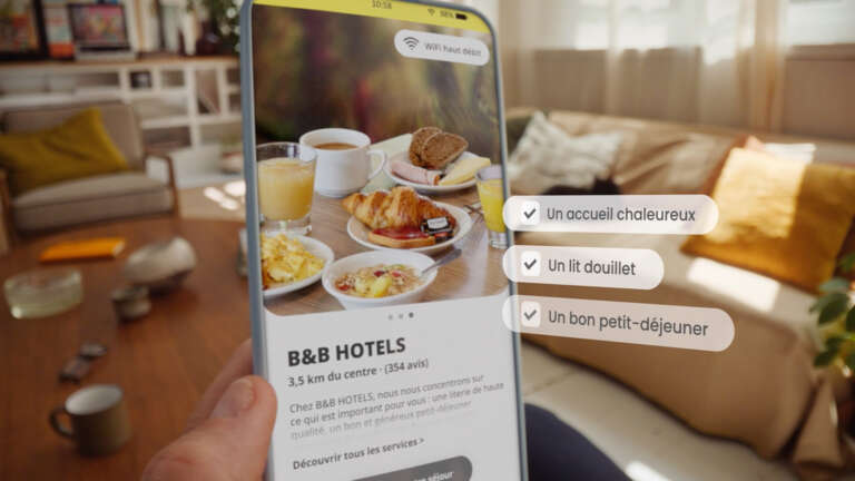 Main tenant un smartphone affichant une page B&B HOTELS avec un petit-déjeuner appétissant et les critères : "Un accueil chaleureux", "Un lit douillet", "Un bon petit-déjeuner".