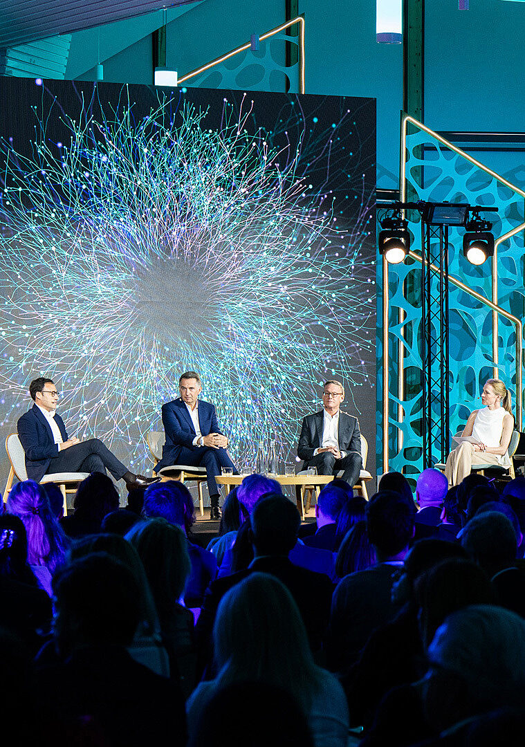 Plenary conference with four speakers seated on stage in front of a giant screen with dynamic light patterns.