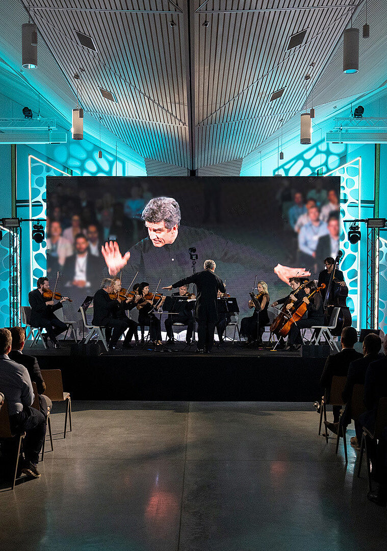A conductor leads a string ensemble on stage, with his performance broadcast on a large screen behind him.